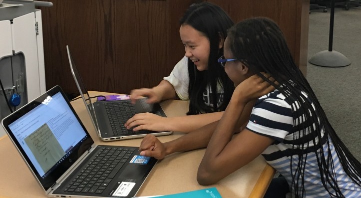 two students working at laptops and putting their heads together to decode historic documents. Both are smiling.